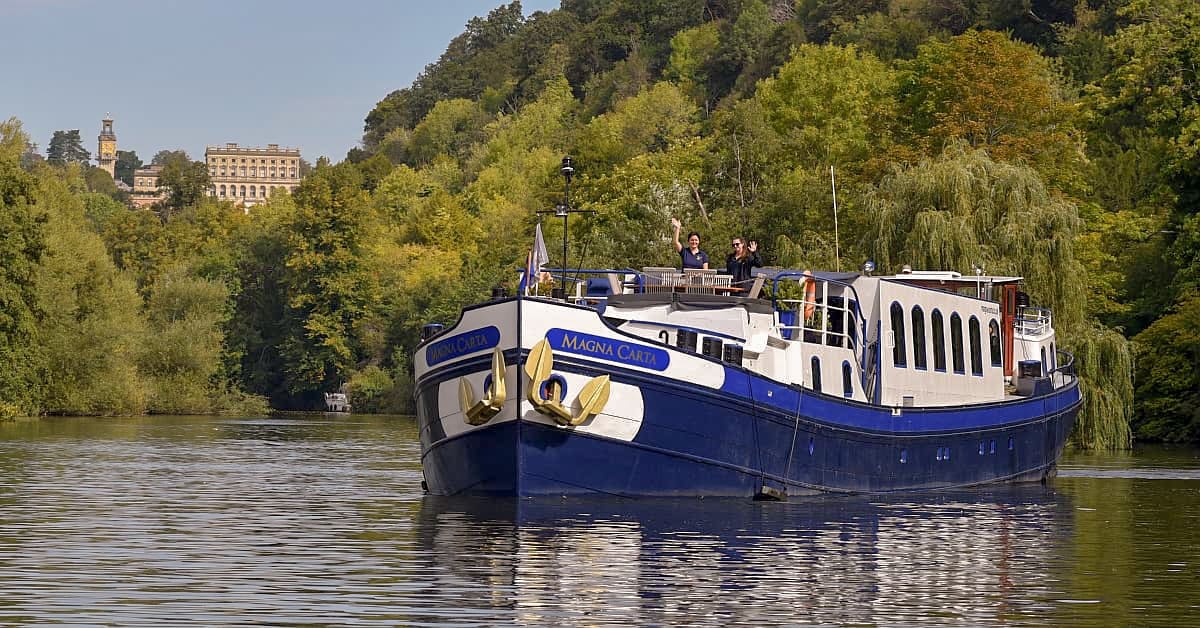 A luxury river cruise barge named Magna Carta navigates a picturesque European river surrounded