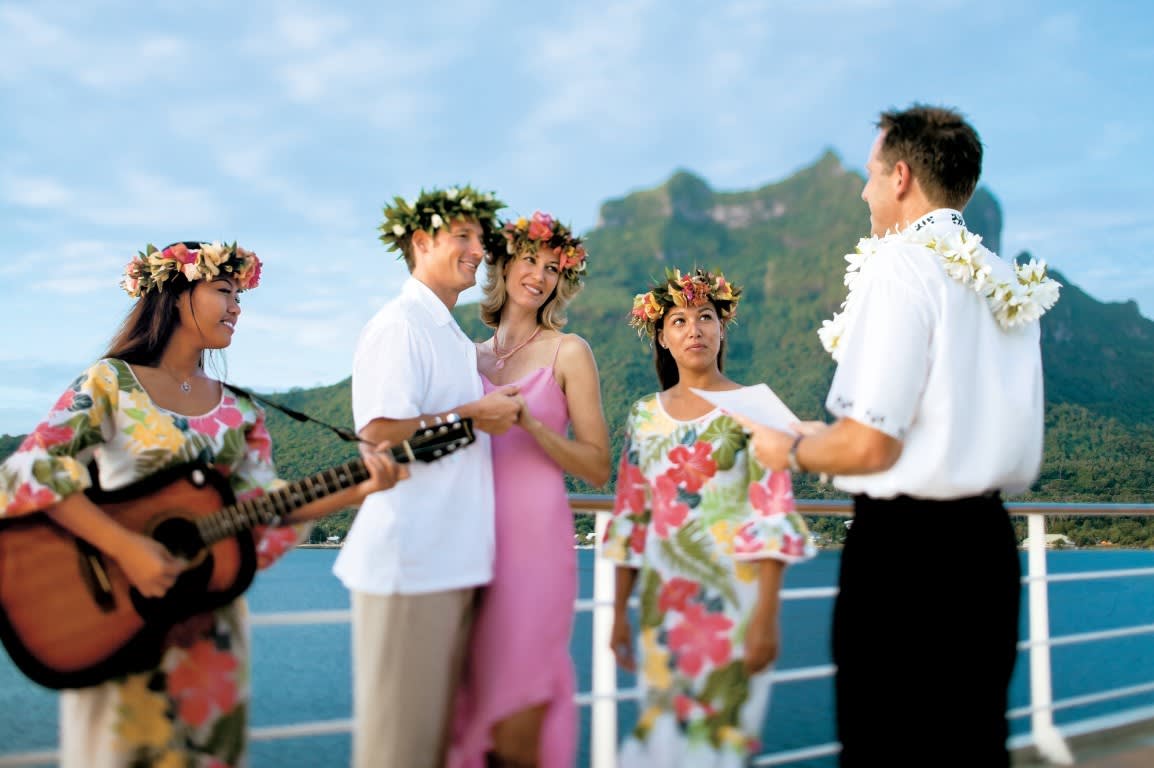 A joyful wedding celebration on a cruise ship's deck featuring five people in tropical attire and flower garlands, with a lush green volcanic island and turquoise ocean visible in the background. The scene captures a Hawaiian or South Pacific-themed destination wedding moment.