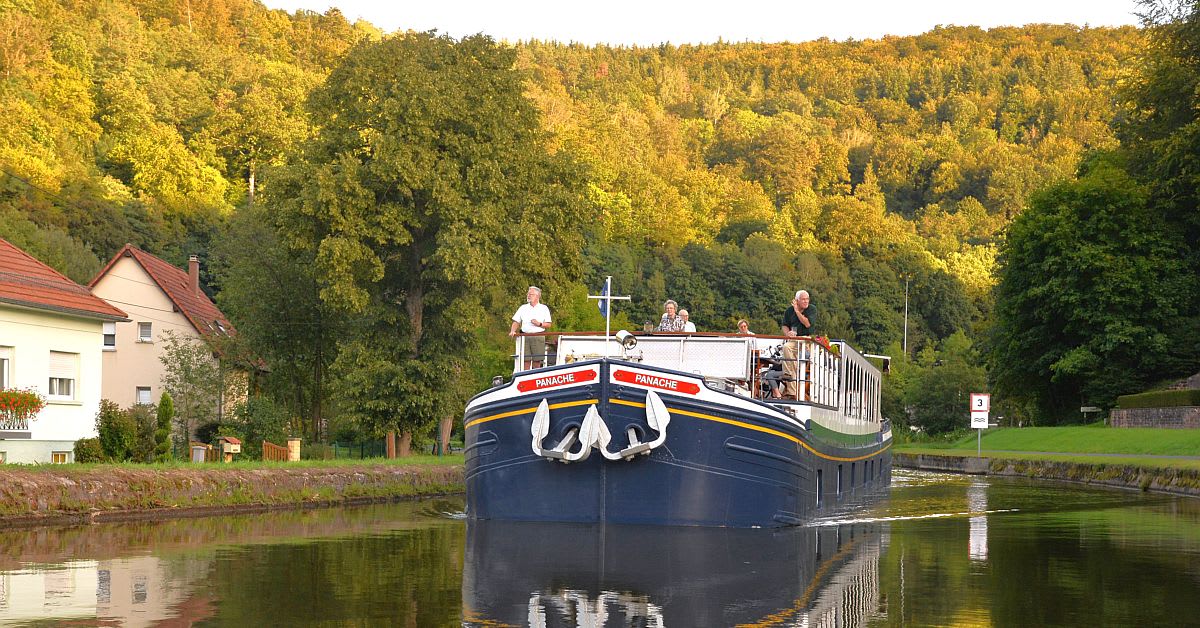A traditional blue river barge named 'Panache' navigates a calm European canal with passengers