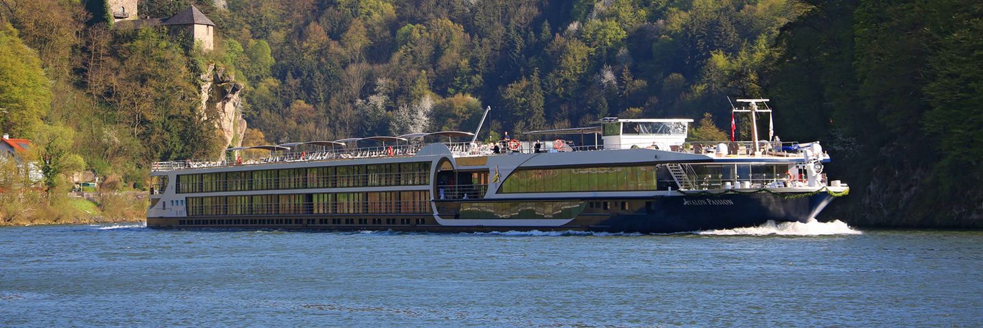 A modern river cruise ship navigates a scenic European river corridor flanked by forested cliffs