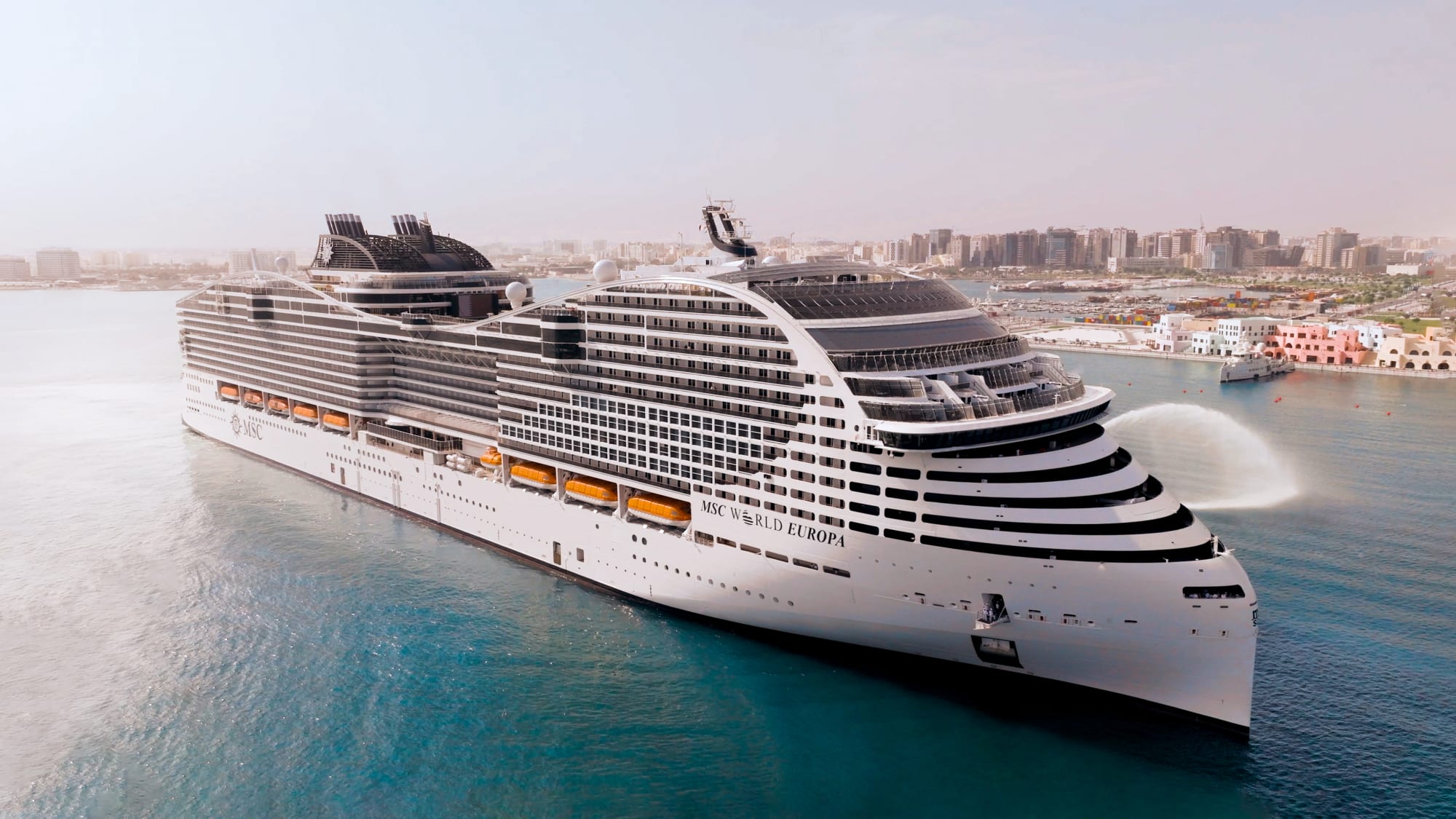 A large modern cruise ship, the MSC World Europa, is anchored in turquoise waters near a Mediterranean coastal city with white and coral-colored waterfront buildings visible in the background. The vessel features a distinctive white hull with multiple deck levels, black and white striping on the superstructure, and large funnel stacks characteristic of contemporary mega-ship design.