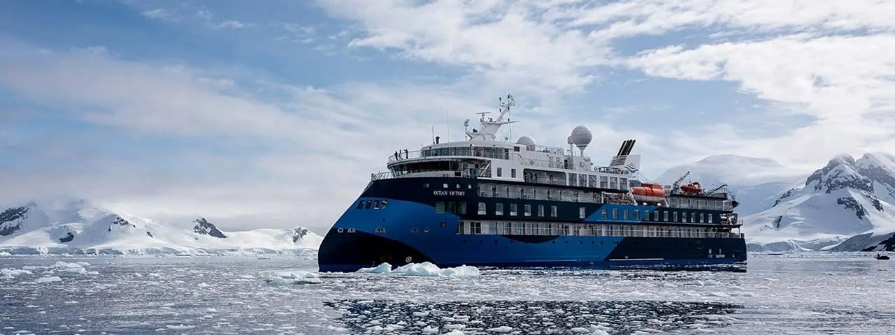 A modern expedition cruise ship with distinctive blue and black hull navigates through icy Antarctic waters surrounded by snow-capped mountains and floating ice. The vessel is equipped with multiple decks, advanced navigation systems, and lifeboat stations, designed specifically for polar exploration cruising.