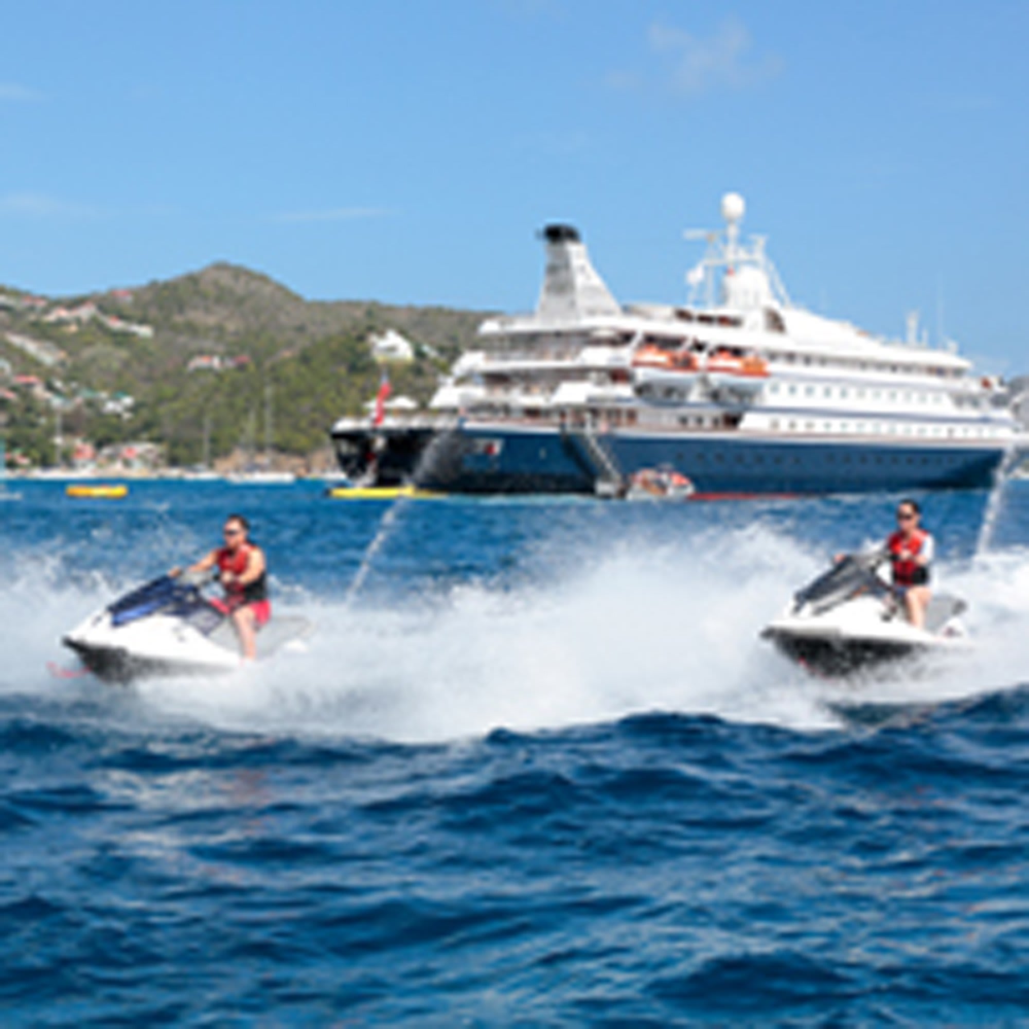 Two jet skiers in red life vests enjoy water sports in front of a large cruise ship anchored in