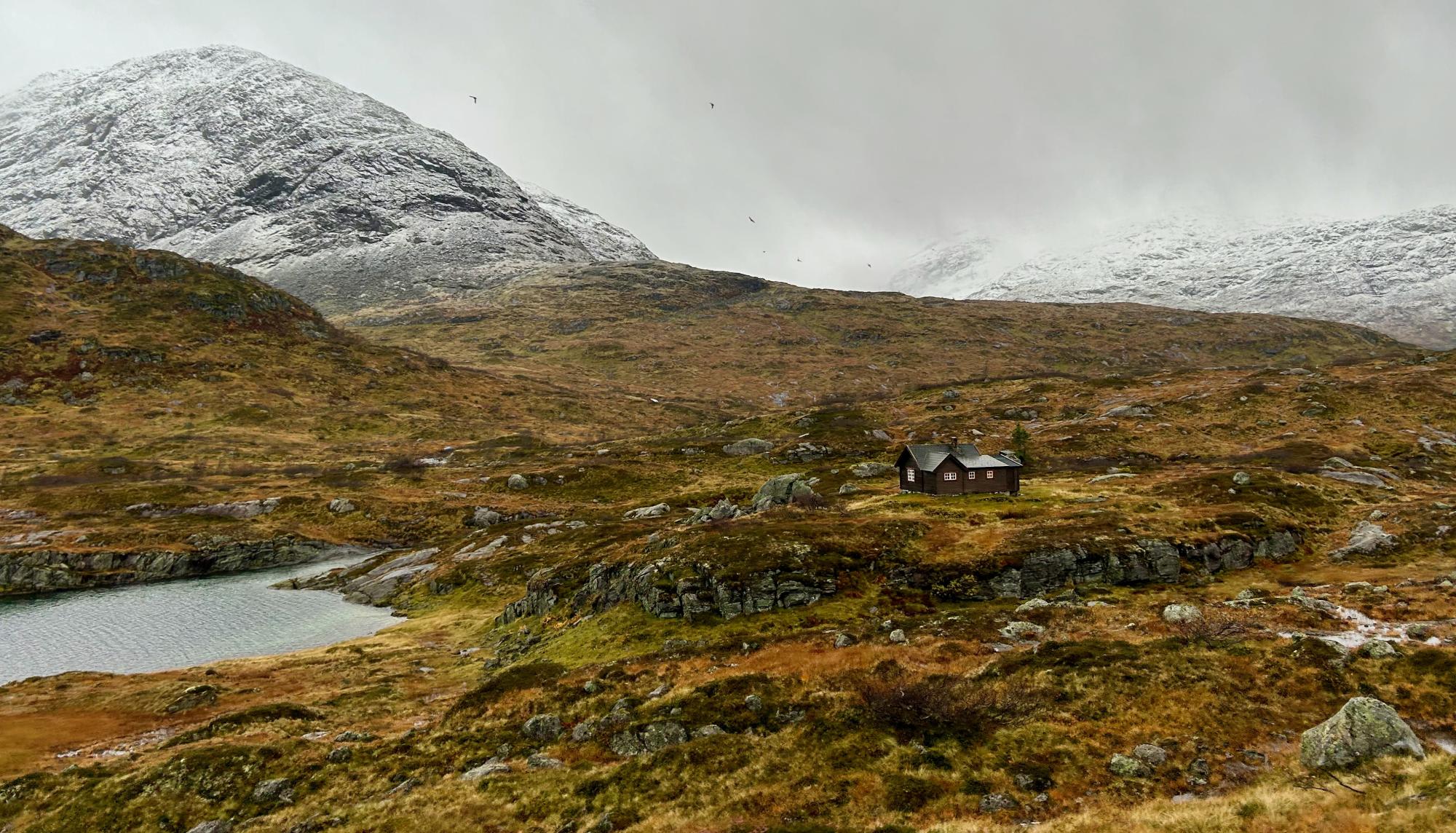 A solitary red wooden cabin sits in a remote, tundra-covered valley beneath snow-capped mountains, with a small glacial pond in the foreground and sparse vegetation characteristic of Arctic or subarctic terrain.