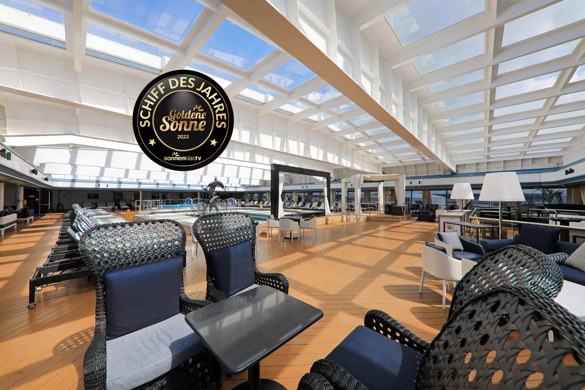 A contemporary indoor aquatic complex featuring a large swimming pool, modern black mesh loungers with blue cushions, and an expansive skylight ceiling that floods the space with natural sunlight. The sophisticated design includes wooden flooring, casual seating areas, and panoramic windows overlooking outdoor facilities, earning recognition as the Ship of the Year (Schiff des Jahres) in 2023.