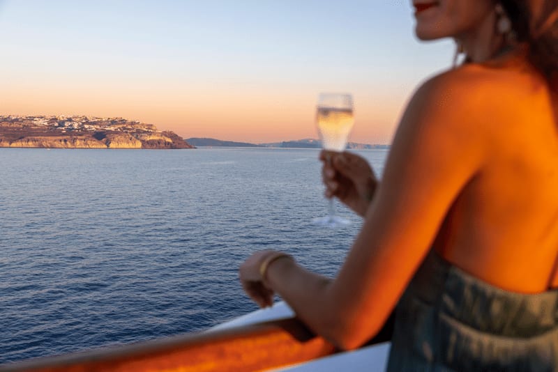A passenger in an orange shirt holds a glass of champagne while gazing at a stunning sunset over