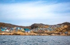 A picturesque coastal settlement nestled against arid, rocky hills viewed from the water, featuring colorful buildings clustered along the shoreline with a calm sea in the foreground and blue sky above. The architecture and landscape suggest a Mediterranean or Middle Eastern location with a dry, semi-arid climate.