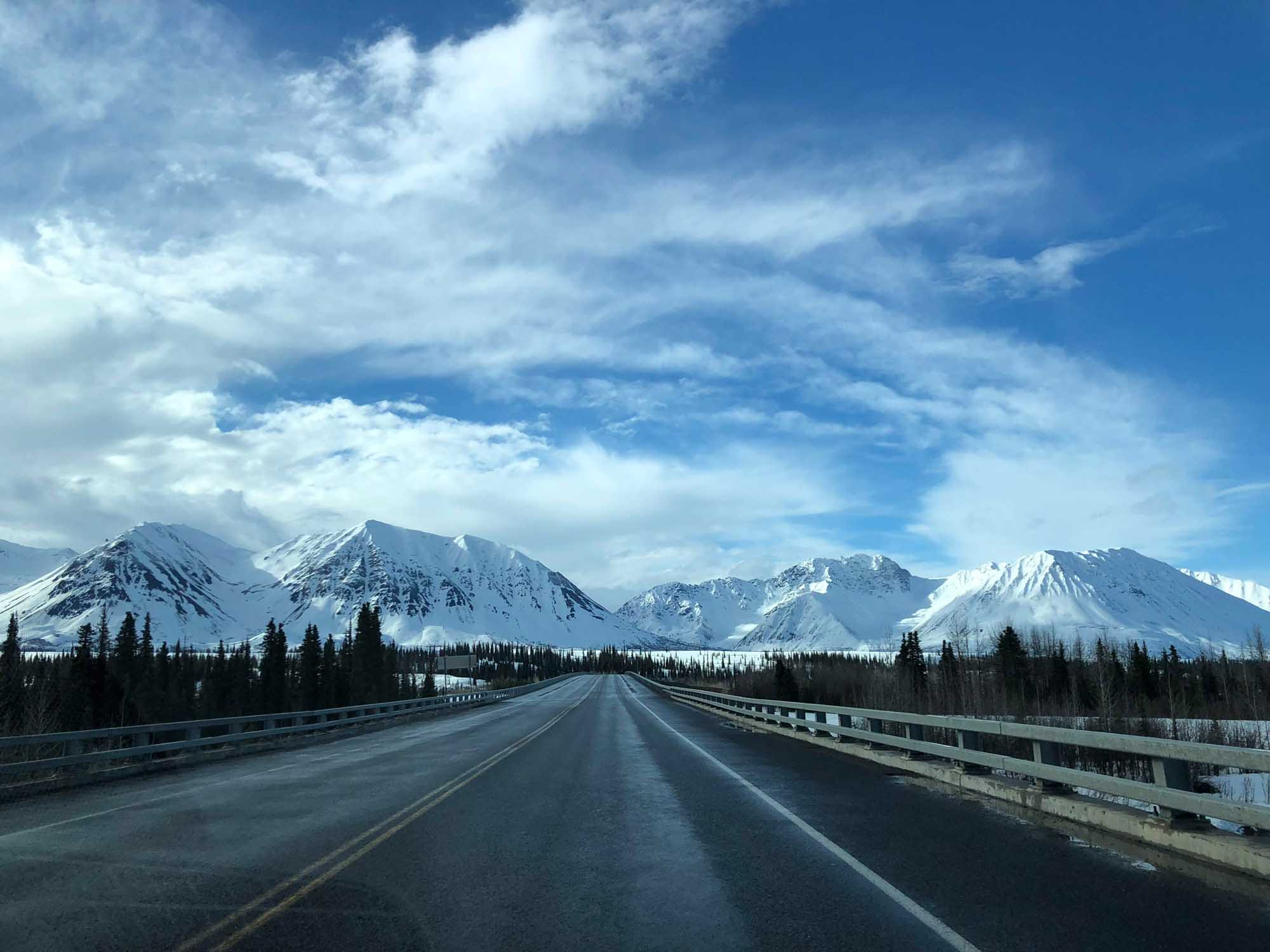 A straight highway stretches toward a dramatic range of snow-covered volcanic peaks under a brilliant blue sky with wispy clouds. Dense coniferous forests line both sides of the road, with metal guardrails visible along the bridge or elevated section, creating a striking perspective toward the distant mountains.