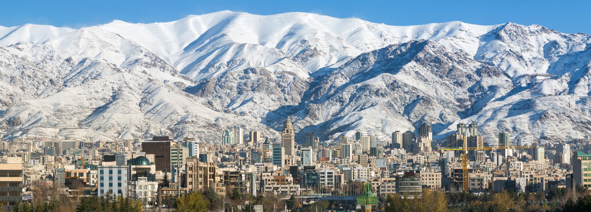 A stunning urban landscape showing the dense cityscape of Tehran, Iran, dominated by numerous high-rise buildings and traditional architecture, set against the dramatic backdrop of the snow-covered Alborz mountain range under a clear blue sky.