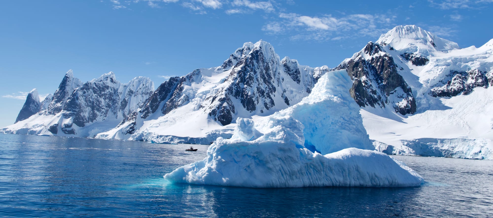 A massive blue-white iceberg floating in the foreground with dramatic snow-covered mountains