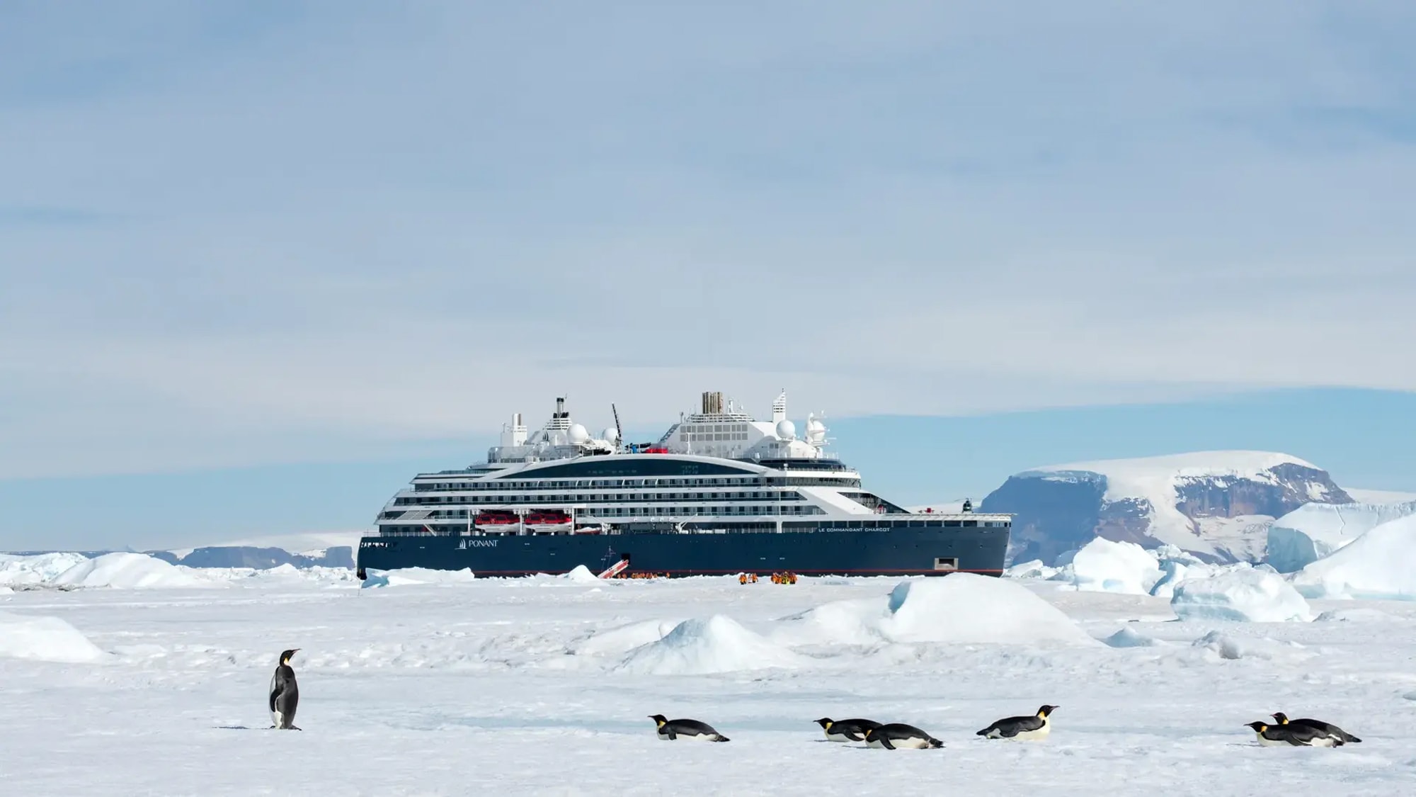 A sleek, modern cruise ship navigates through calm waters with rugged mountain terrain in the background. The vessel is adorned with multiple satellite domes and has a clean, contemporary design.