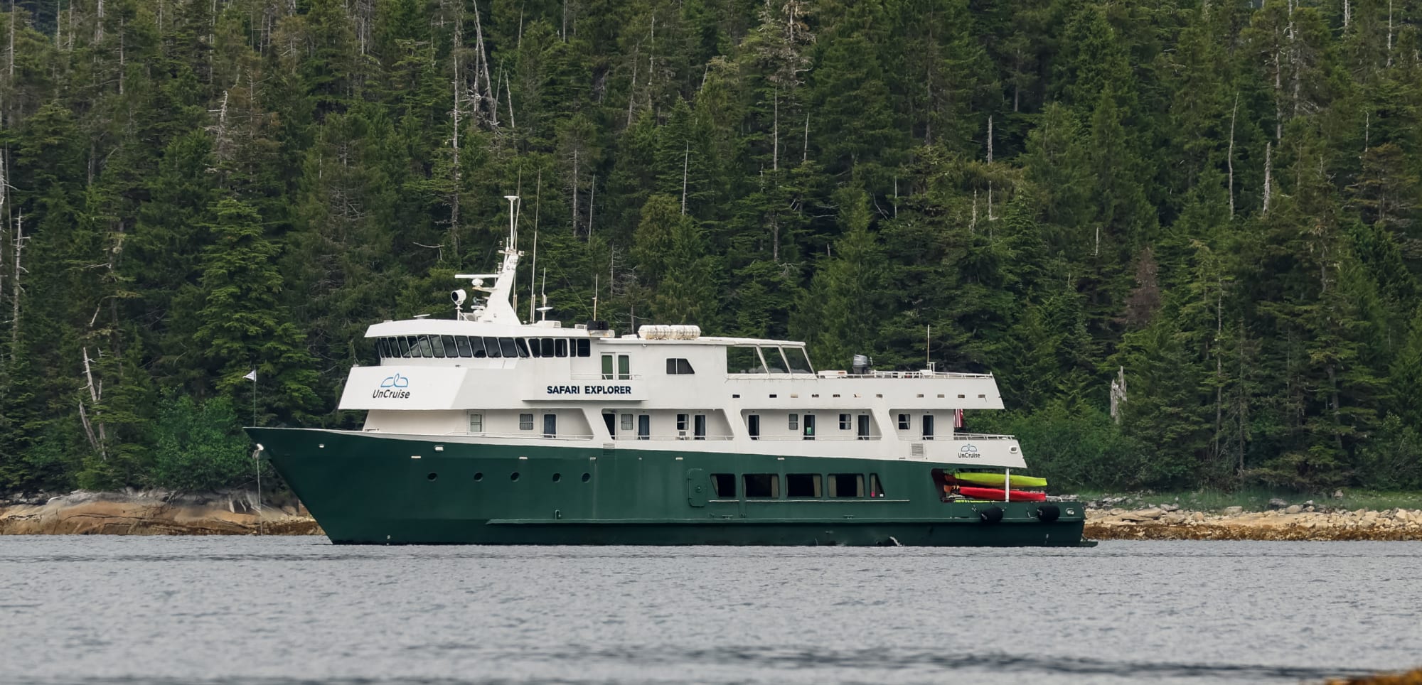 A green-hulled expedition cruise ship named Safari Explorer anchored near a rocky shoreline