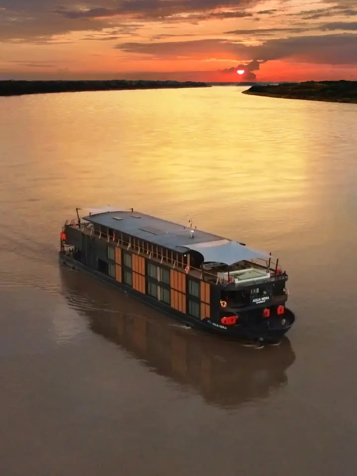 A modern river cruise vessel with distinctive orange and dark hull coloring navigates a calm, wide