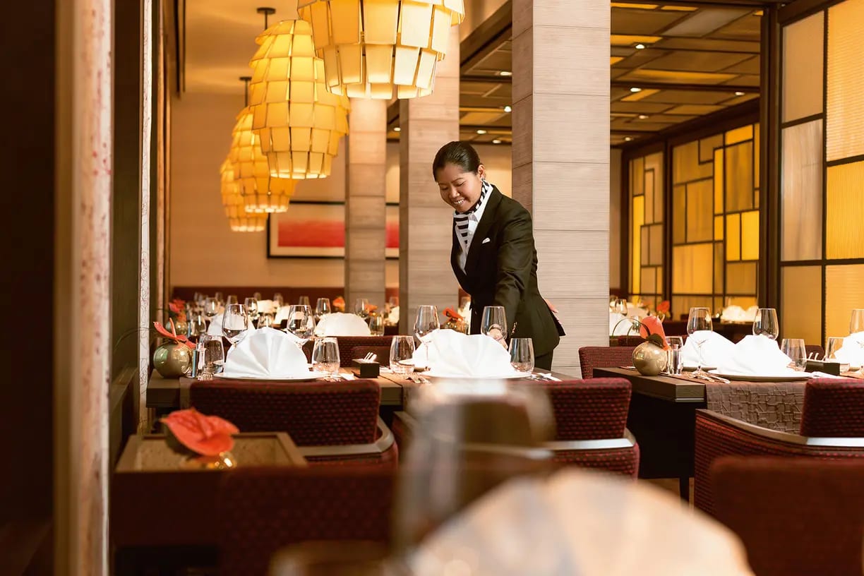 A professional server prepares an elegant dining table in a high-end restaurant with warm golden lighting, white linens, and sophisticated place settings. The sophisticated interior features modern pendant lights, floor-to-ceiling windows, and upscale furnishings typical of luxury cruise ship or hotel dining venues.