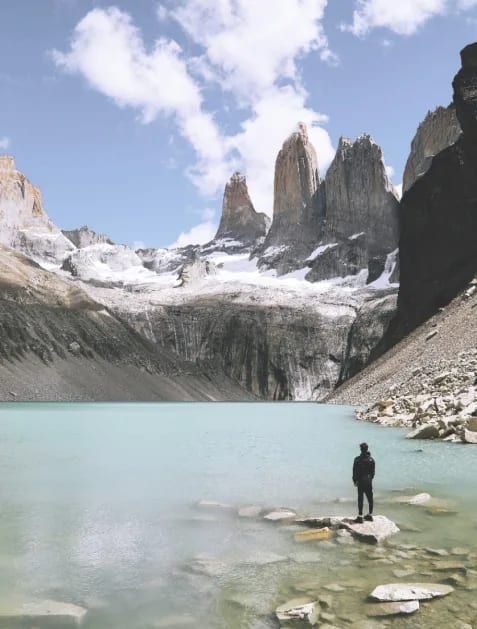 A solitary hiker stands on rocky shore beside a pristine turquoise glacial lake, gazing up