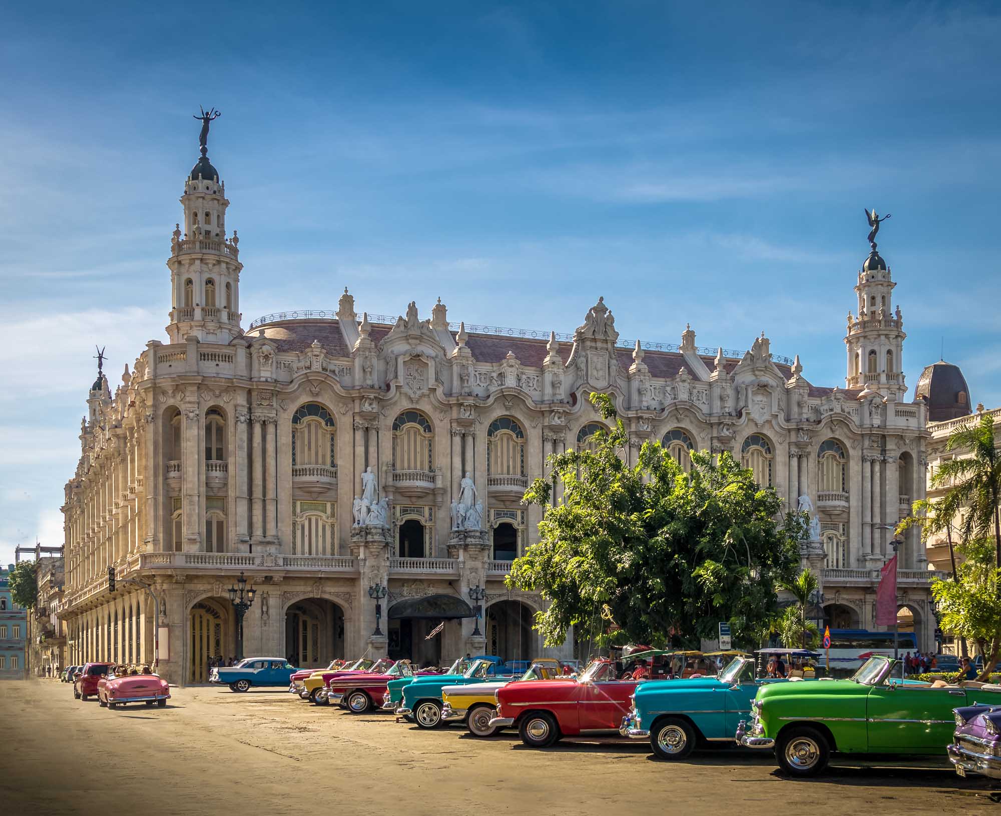 A vibrant collection of colorful 1950s American classic cars parked in front of the ornate Gran Teatro de La Habana, a stunning Baroque Revival theater building in Cuba's capital city. The scene captures the iconic street culture of Havana with its distinctive pastel-colored vintage automobiles and grand historic architecture.