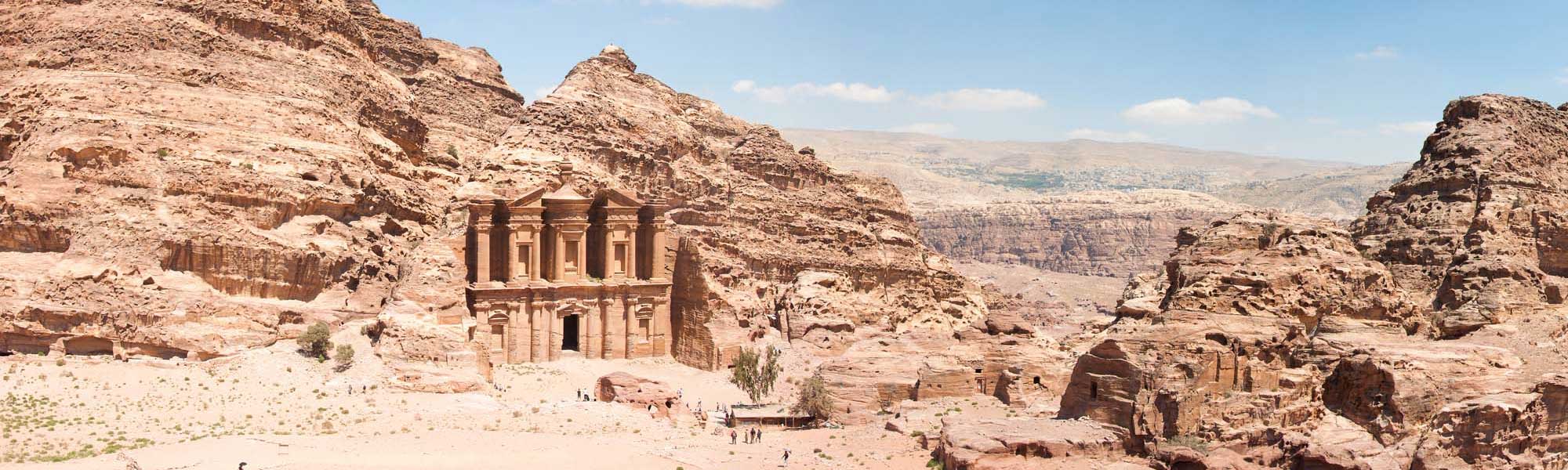 An iconic view of Al-Deir (The Monastery), one of Petra's most magnificent rock-carved facades, featuring grand Nabatean columns and architectural details set within towering sandstone cliffs in the Jordanian desert. The massive structure stands as a testament to ancient engineering carved directly from the russet and tan stone formations.