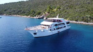 A modern multi-deck cruise ship anchored in a pristine turquoise Mediterranean cove surrounded