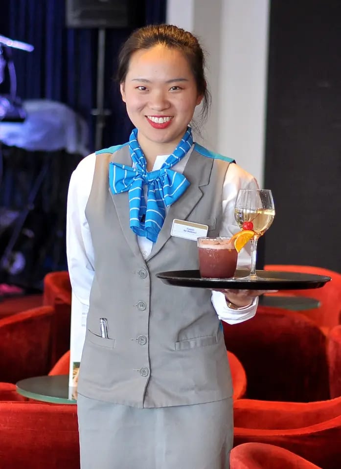 A smiling crew member in Celestyal Cruises uniform (gray vest with blue scarf) stands in an elegant