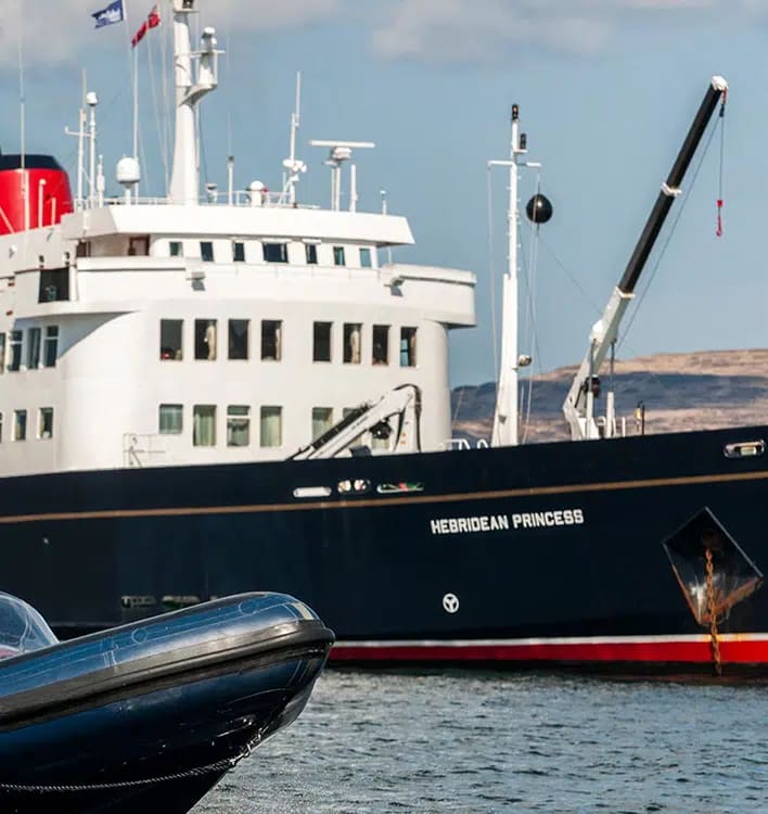 The Hebridean Princess, a small luxury cruise ship, is moored at a Scottish port with its distinctive white superstructure and black hull clearly visible. A small tender boat is secured alongside the vessel, with rolling hills visible in the background under a partly cloudy sky.
