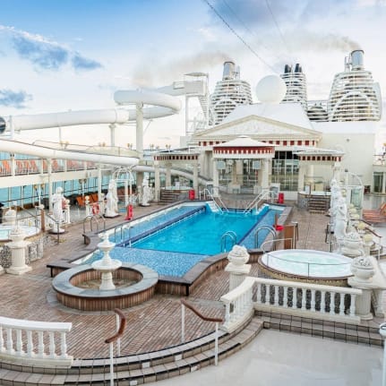 An expansive cruise ship pool deck featuring a large turquoise swimming pool, hot tubs, water