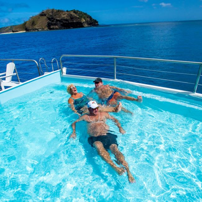 A family enjoys swimming and relaxing in a turquoise pool deck aboard a cruise ship, with pristine tropical waters and a lush island visible in the background under clear blue skies.