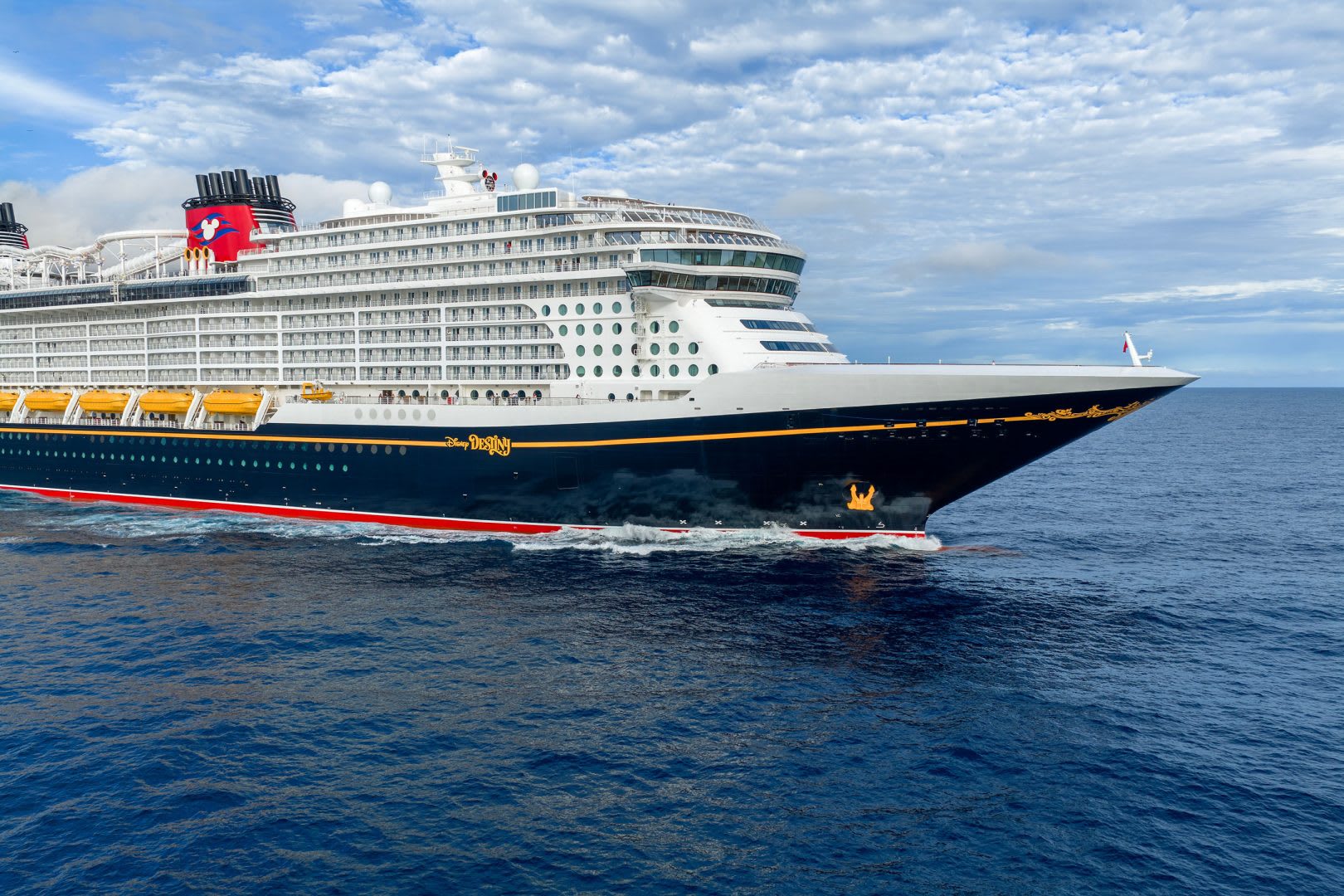 A massive modern cruise ship with distinctive Disney branding on its red funnel featuring Mickey Mouse, anchored in calm blue waters under a partly cloudy sky. The vessel displays the characteristic white superstructure and dark hull typical of contemporary mega-ships.