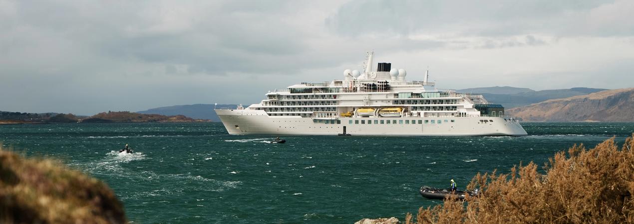 A white luxury cruise ship anchored in a turquoise ocean with rocky coastline and mountains in