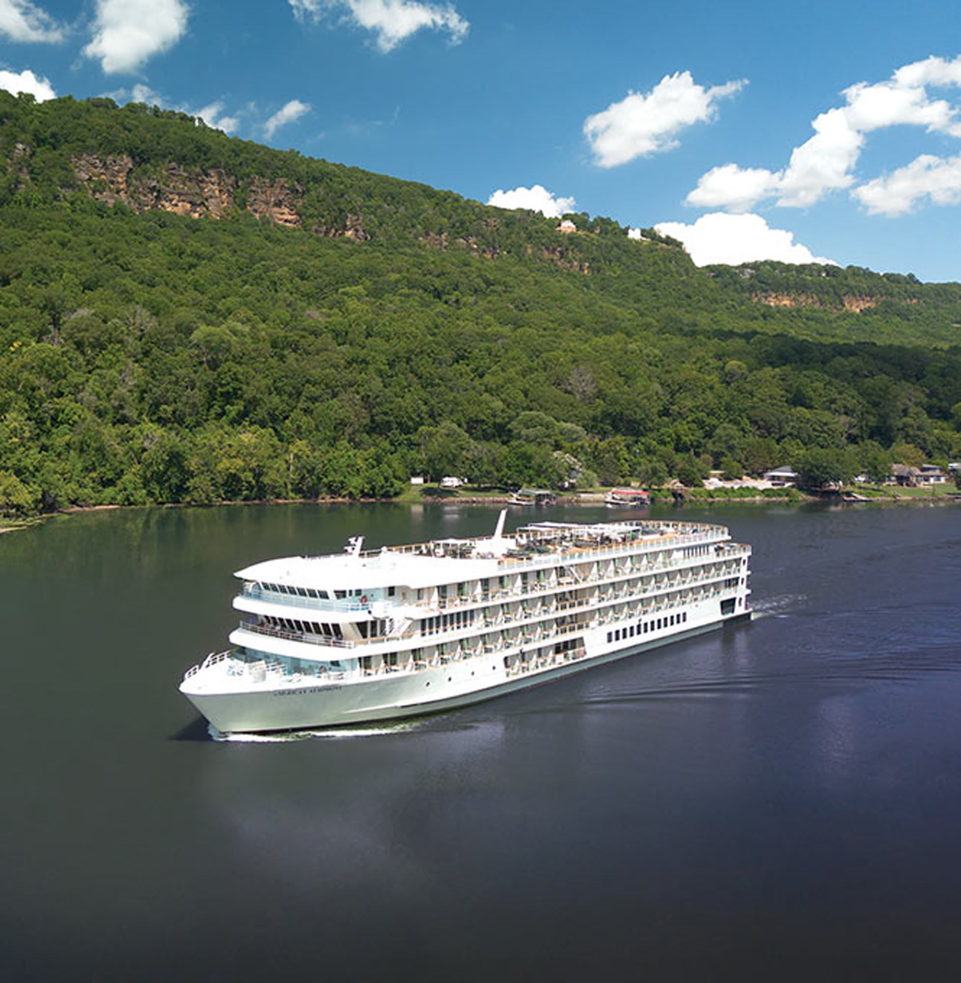 A large white river cruise ship navigates a calm river flanked by lush, densely forested hillsides
