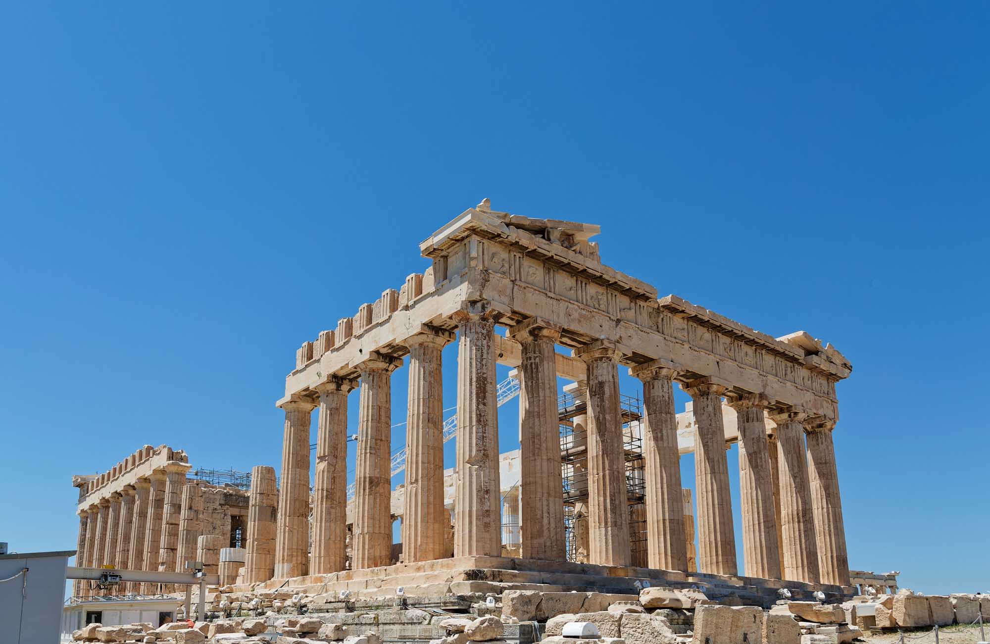 The iconic Parthenon temple stands majestically on the Athenian Acropolis against a clear blue sky. The ancient Greek structure features its distinctive Doric columns and ornate architectural details, with visible restoration scaffolding indicating ongoing preservation work.