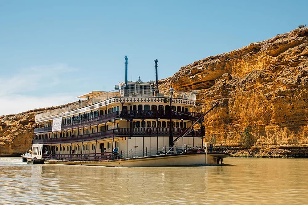 A classic multi-deck paddlewheel riverboat with distinctive twin smokestacks navigates a muddy river, with dramatic golden-hued limestone cliffs towering in the background. The vessel features traditional stern-wheel design with ornate white railings and multiple passenger decks typical of Mississippi River-style cruising.