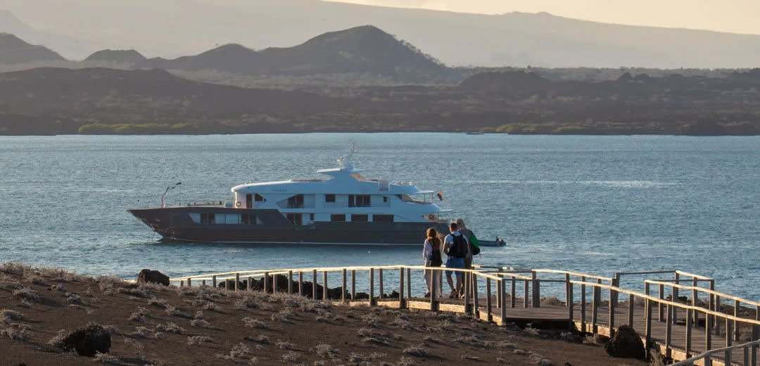 A blue and black expedition cruise ship anchors offshore at a pristine Galápagos island beach