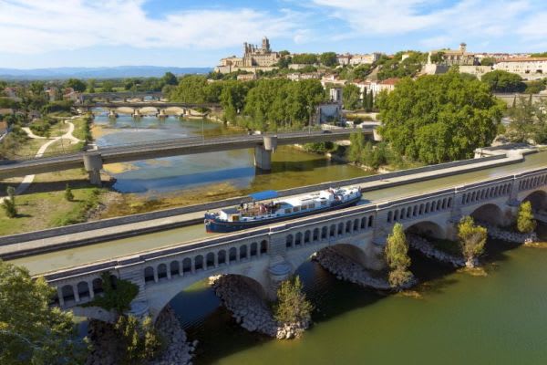 A modern river barge travels along the Lot River in southwestern France, passing beneath a historic