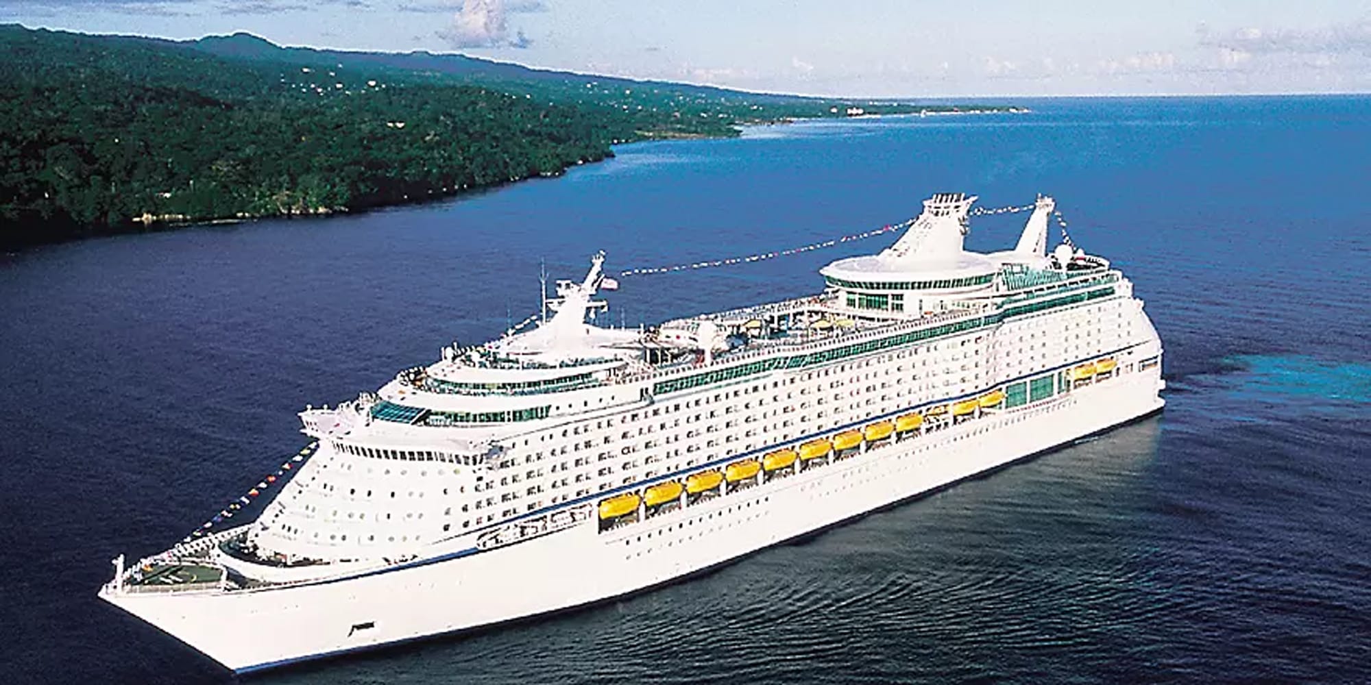 A massive modern cruise ship with white hull and distinctive yellow lifeboats is anchored in clear