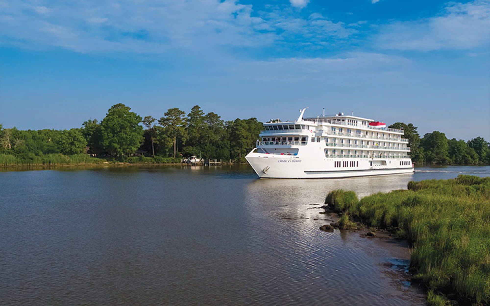 A large multi-deck river cruise ship navigates a calm inland waterway lined with marsh grasses