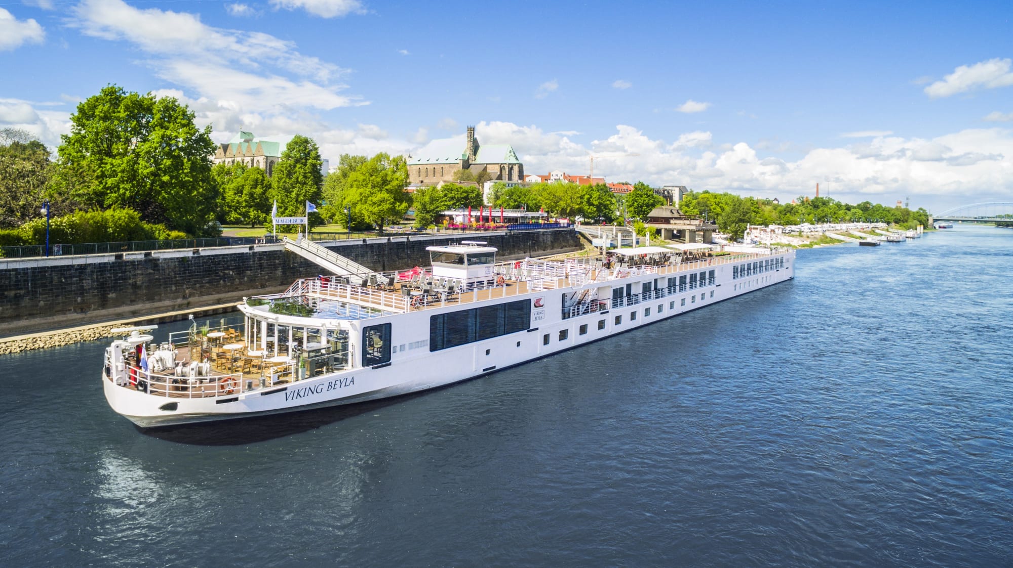 A white Viking river cruise ship docked near a historic riverside city with green trees and architectural landmarks. The vessel is moored in a lock with a picturesque urban landscape in the background.
