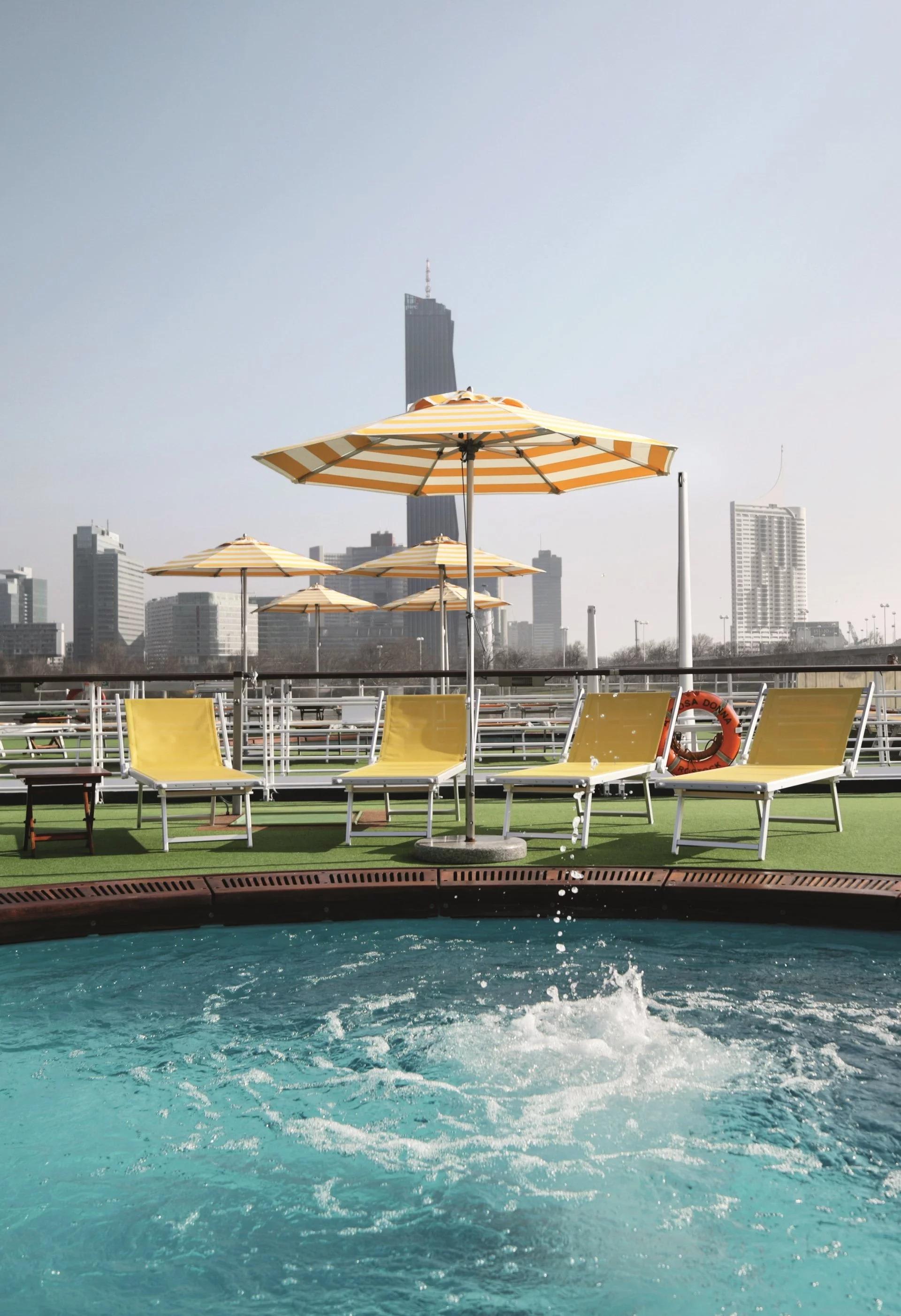 A luxurious riverside pool deck aboard an A-ROSA cruise ship, featuring turquoise waters, yellow