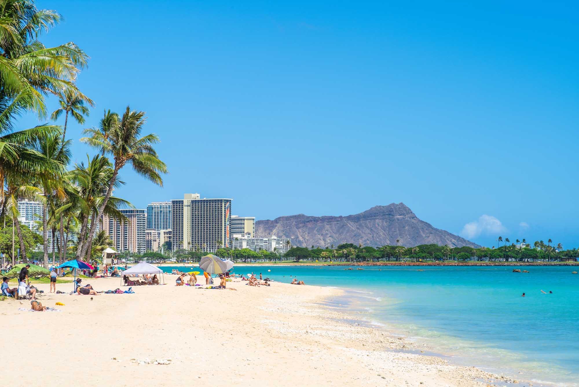 A scenic view of Waikiki Beach in Honolulu, Hawaii, featuring pristine white sand, crystal-clear turquoise waters, and beachgoers enjoying the tropical weather. The iconic Diamond Head mountain rises majestically in the background, with modern high-rise hotels and swaying palm trees framing this quintessential Hawaiian destination.