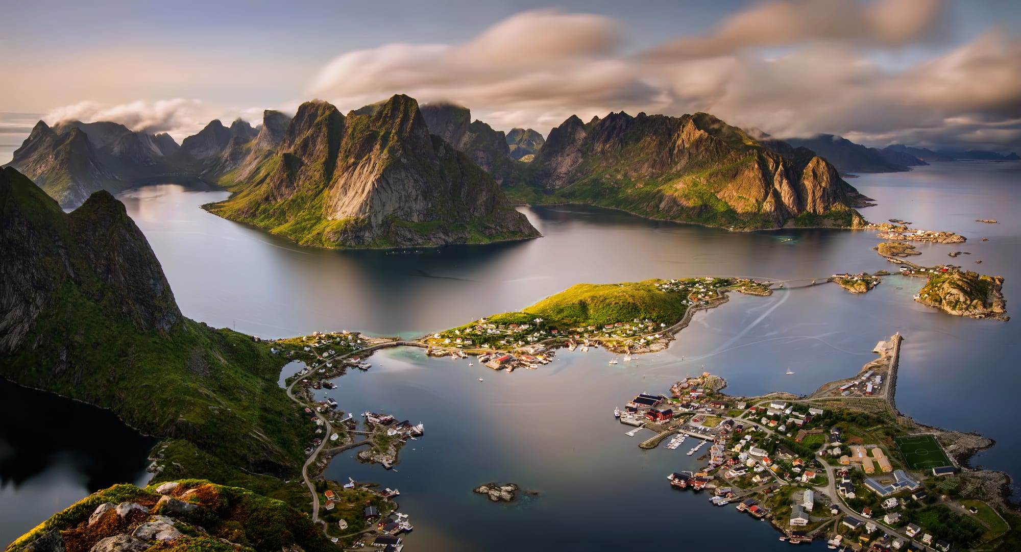 A dramatic aerial photograph of the Lofoten Islands in northern Norway, showcasing towering jagged peaks rising from calm waters, with traditional fishing villages nestled along the coastline. The landscape features steep green-covered mountains, small red and yellow houses, and picturesque harbors surrounded by pristine Arctic waters.
