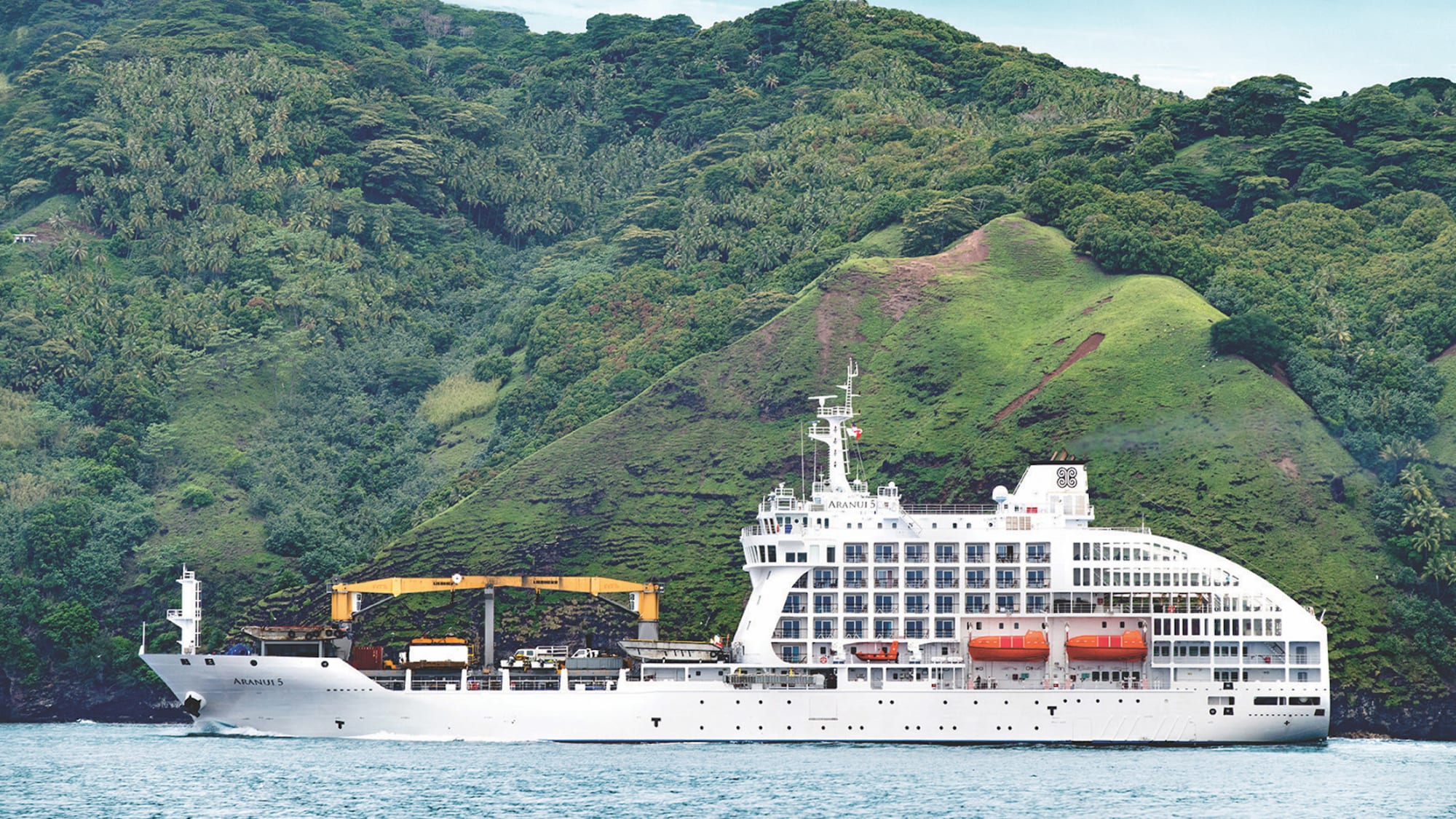 A white cruise ship named Aranui 5 anchored near a densely forested mountainous coastline with green hills and tropical vegetation. The ship appears to be a specialized expedition or cargo-cruise vessel positioned in a scenic maritime setting.