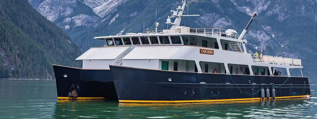 A striking expedition cruise vessel named Cascadia anchored in a pristine glacial fjord surrounded by snow-capped mountains and dense evergreen forests. The ship features a distinctive two-hull catamaran design with white upper decks and dark navy hull, accented with yellow waterline trim, positioned in turquoise alpine waters.