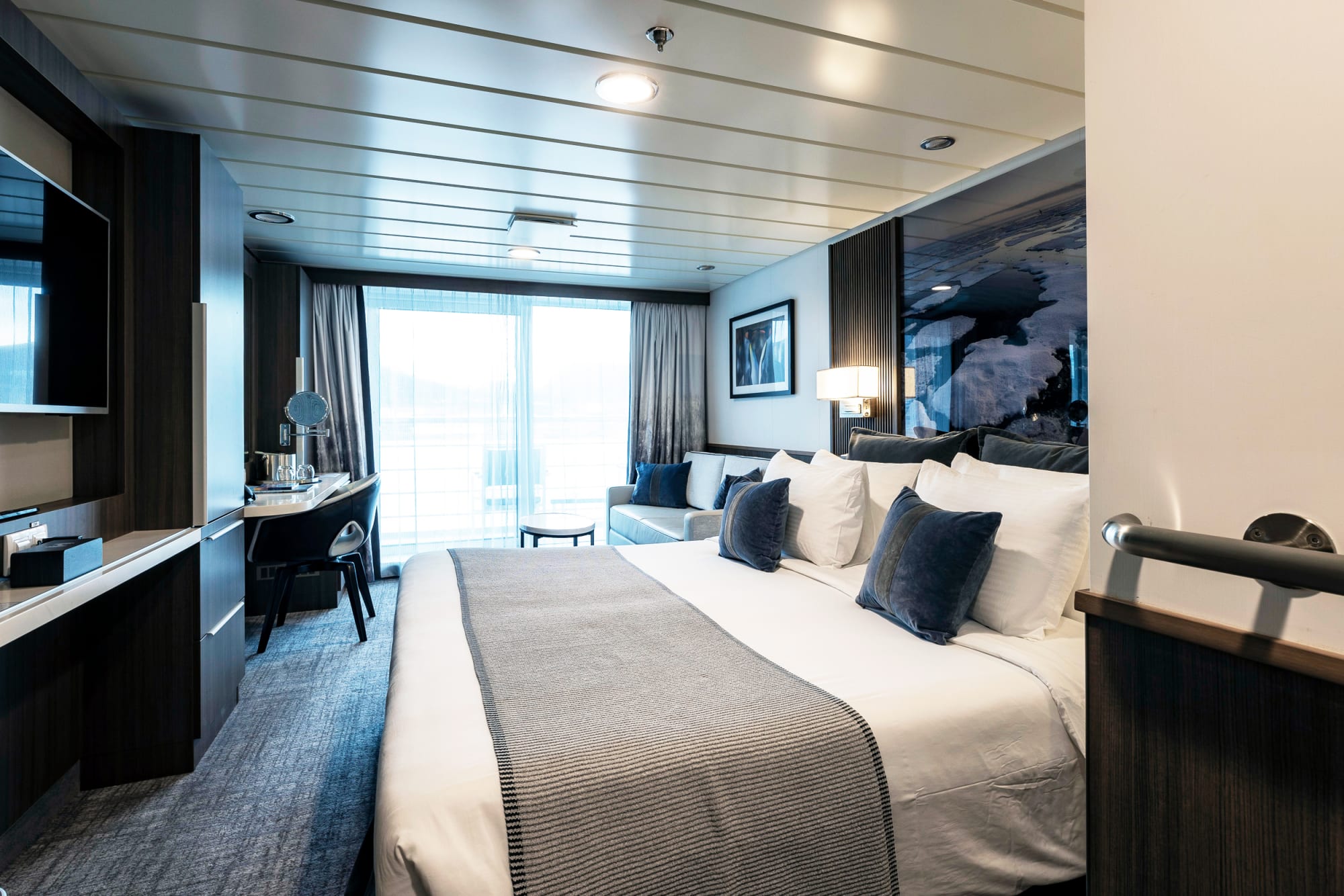 A contemporary luxury cruise ship stateroom featuring a king-size bed with white linens and blue