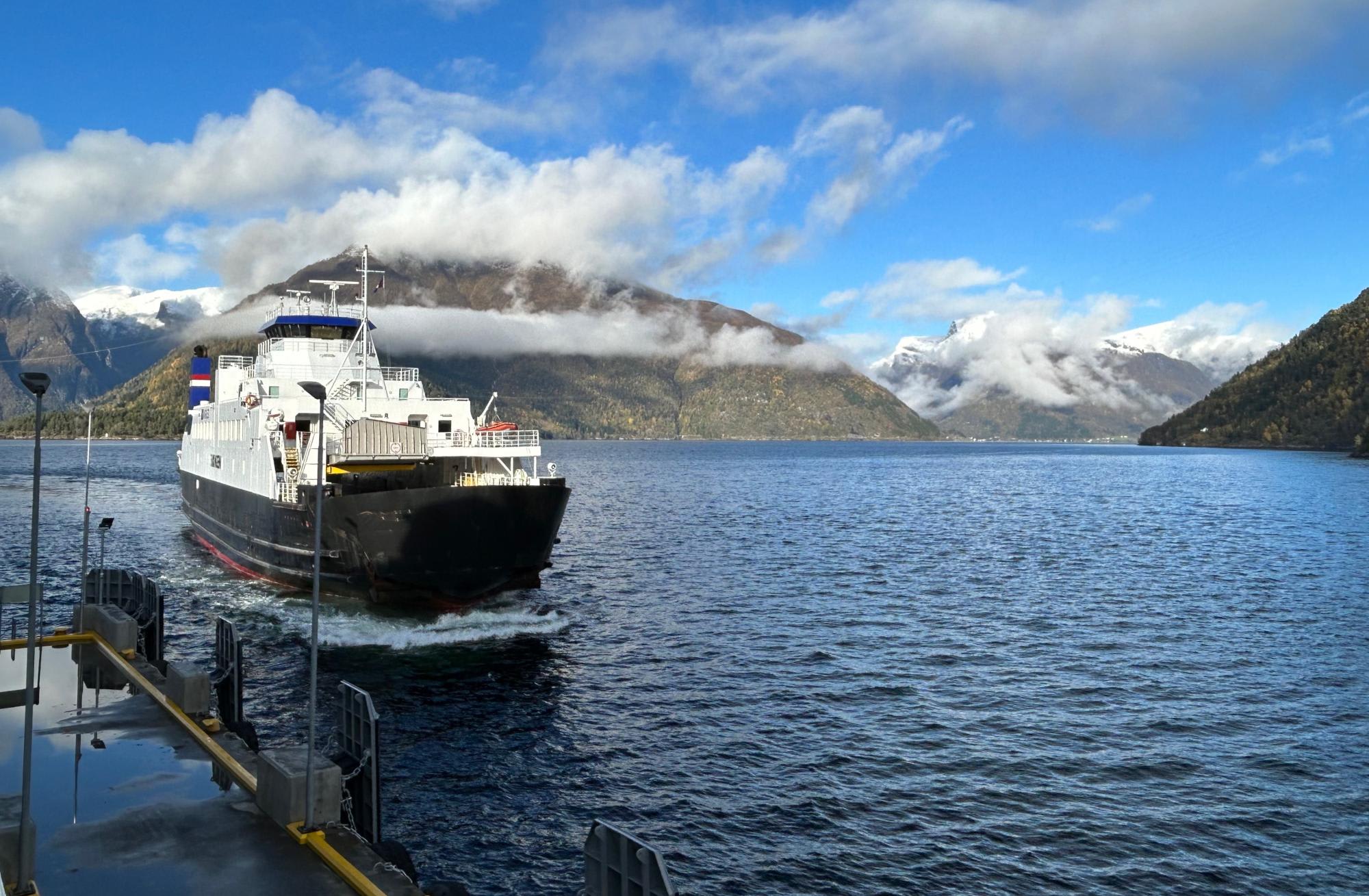 A white and black passenger ferry with Dutch flag markings departs from a dock on a pristine alpine