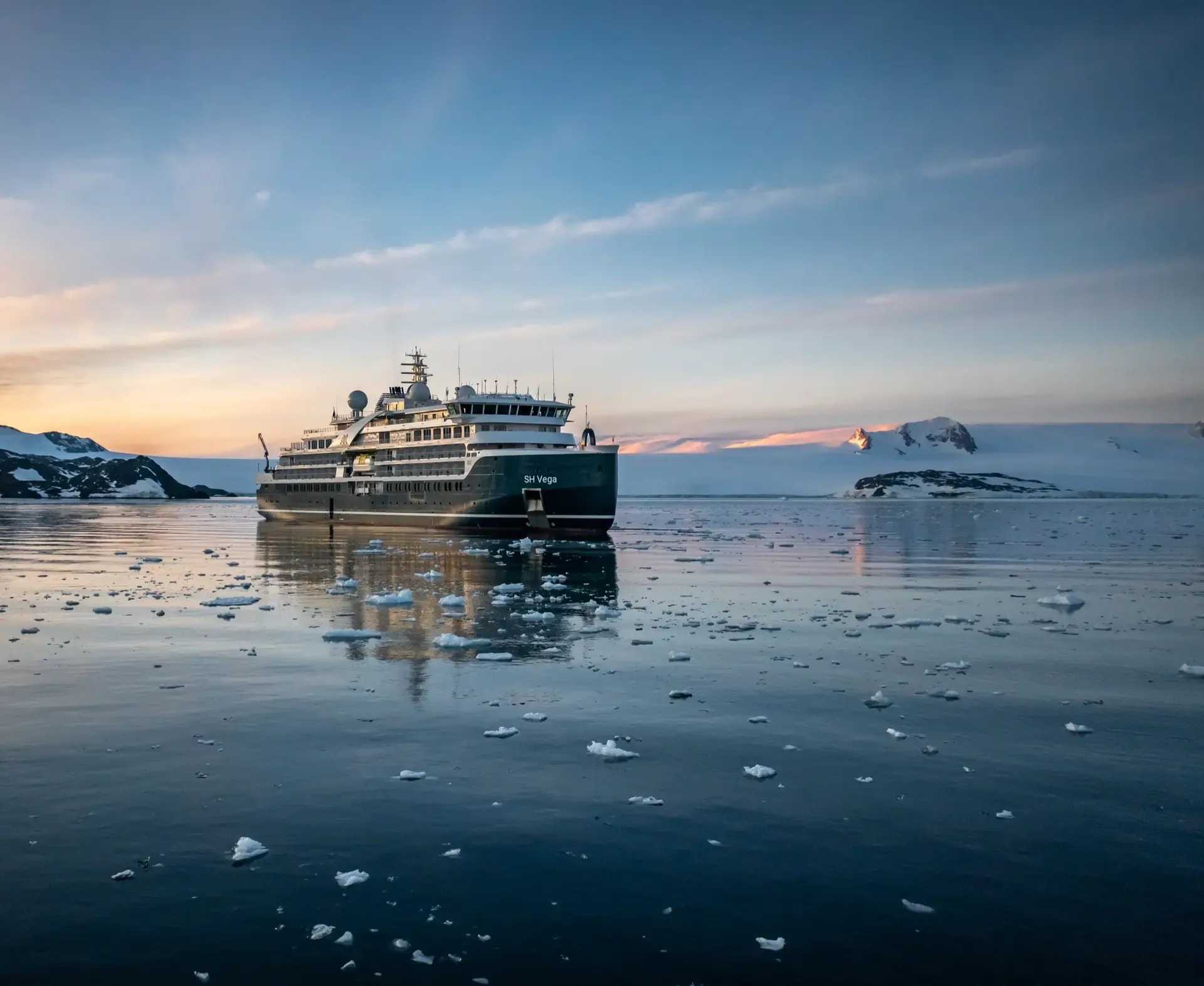 A luxury expedition cruise ship navigates through icy Antarctic waters at golden hour, surrounded