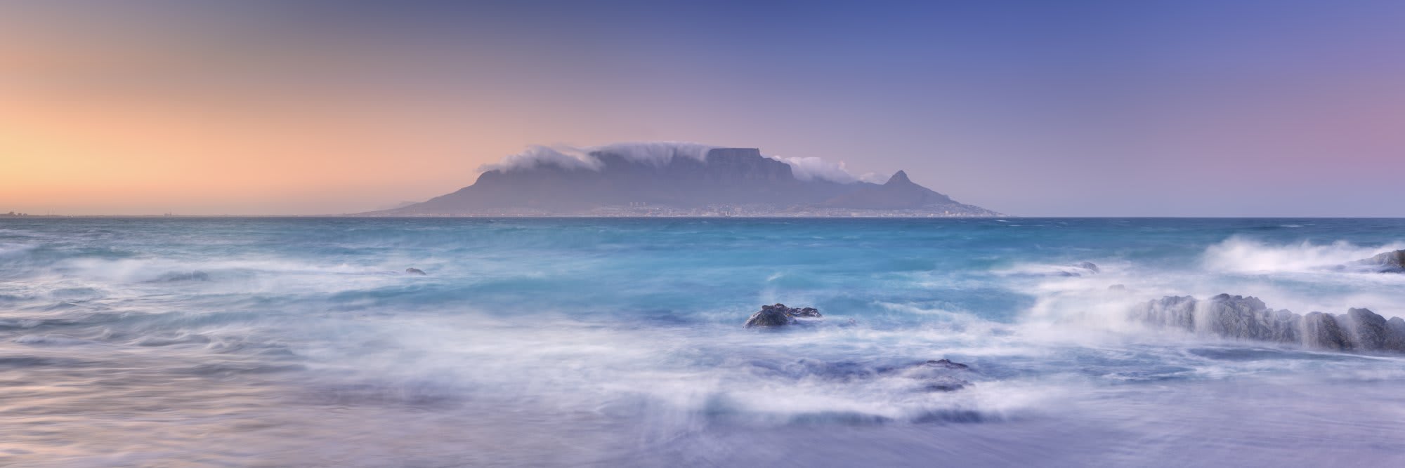 A stunning coastal landscape featuring Table Mountain shrouded in cloud cover at dawn, viewed