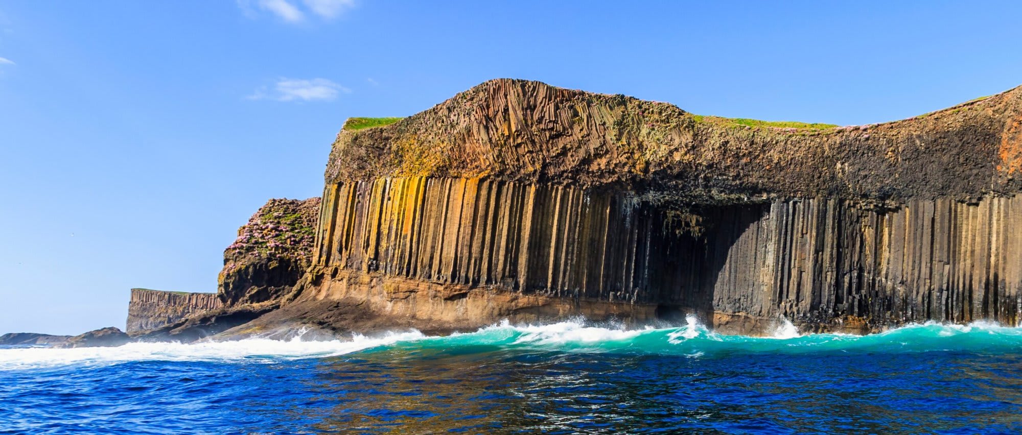 A dramatic coastal landscape featuring distinctive hexagonal basalt columns rising from turquoise waters, with waves crashing against the volcanic rock formations. The scene showcases the UNESCO-recognized geological formations of Staffa in Scotland, a popular destination for cruise ship excursions.