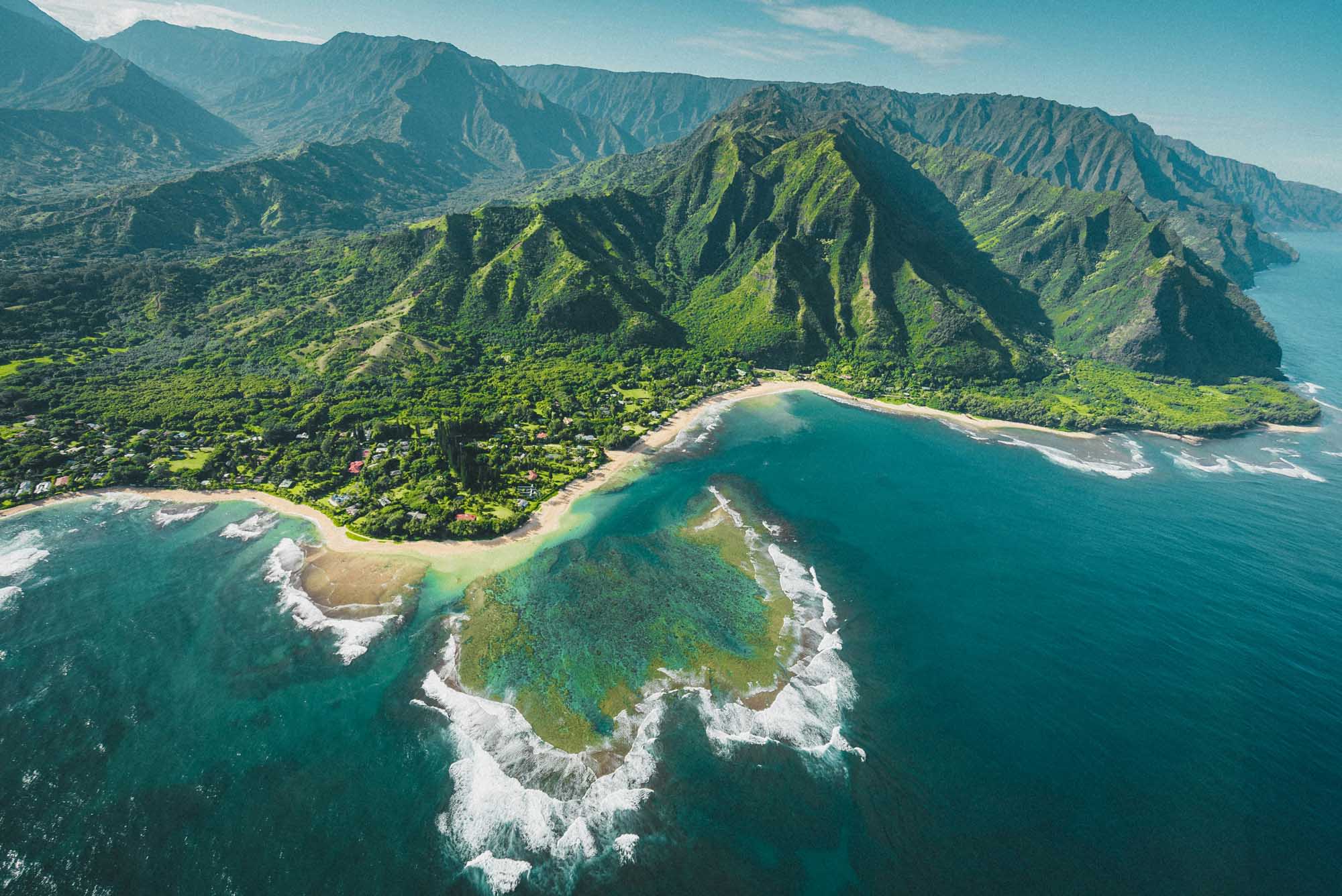 An aerial photograph of the lush Kalalau Valley on Kauai's dramatic Na Pali Coast, featuring verdant cliffs, a pristine sandy beach, turquoise waters, and white-capped ocean waves. The steep fluted pali (cliffs) rise majestically from the valley floor, showcasing the island's stunning volcanic geology and tropical vegetation.