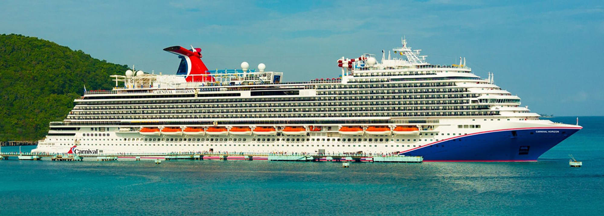A large white and blue Carnival cruise ship docked in a turquoise bay with lush green mountainous