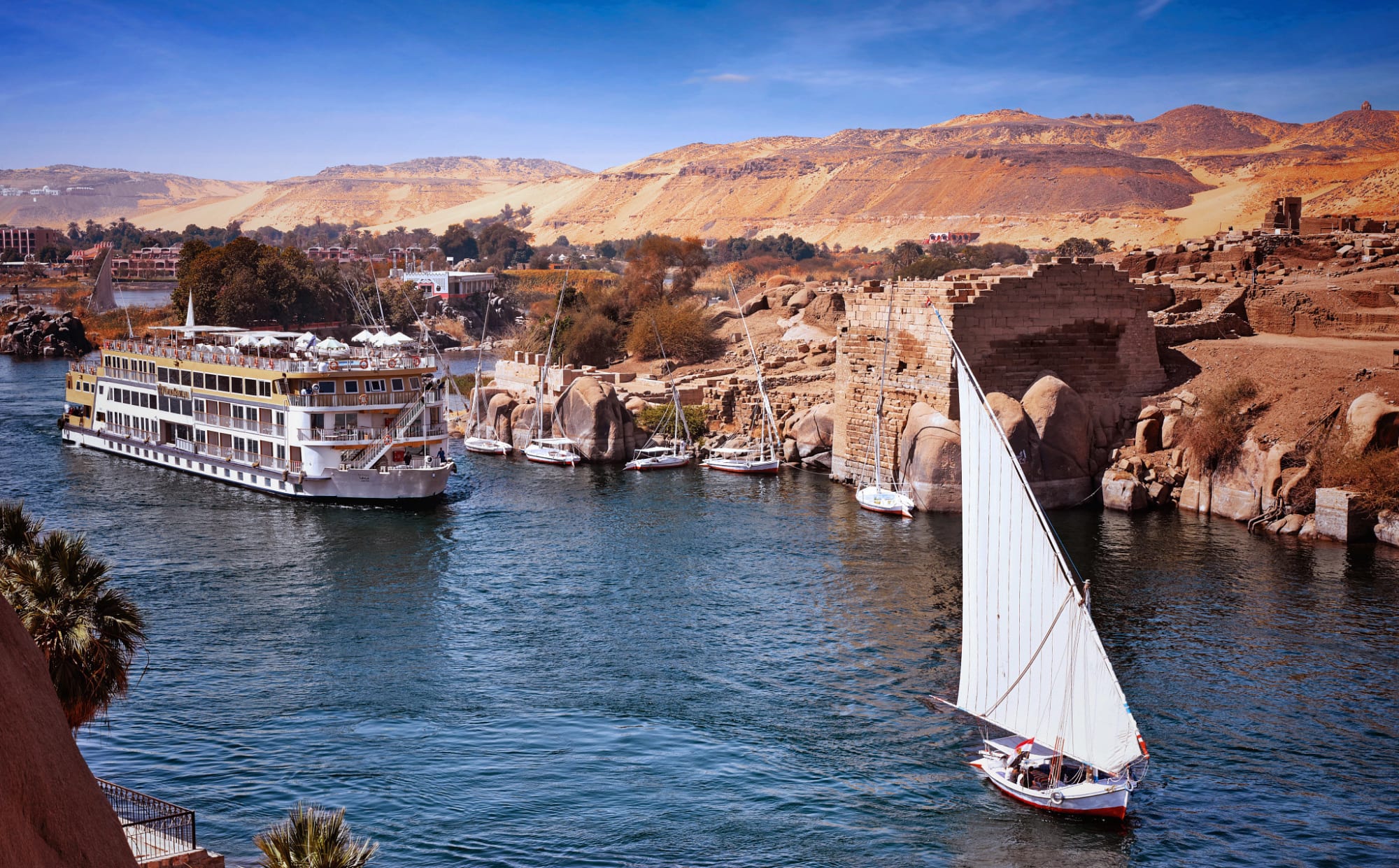 A large river cruise ship docked along the Nile River at Aswan, Egypt, with traditional felucca sailboats and ancient stone ruins visible along the riverbank. The arid desert landscape and palm trees frame the scene under a clear blue sky.