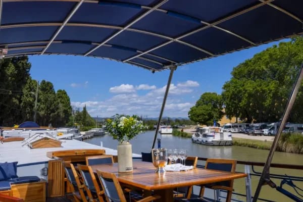 An elegant outdoor dining area situated on a river cruise vessel, featuring a wooden table set for dining under a blue solar panel canopy, with scenic riverside views of moored boats, lush green vegetation, and distant mountains visible beyond the waterway.